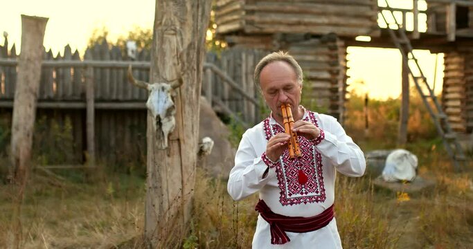 Folklore traditions of the Slavic peoples, a man in a folk costume plays a wooden pipe. Courtyard with a wooden fence and pagan idols, folklore. 4k, ProRes