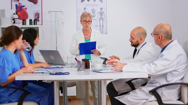 Portrait Of Mature Woman Doctor Heading Committee Meeting At Medical Council Presenting New Medical Procedures Standing At Desk. Group Of Physicians Talking About Symptoms Of Disease In Clinic Office