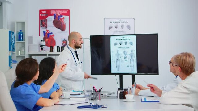 Experienced man doctor analysing human skeleton image together with cvalified colleagues in meeting room, pointing on monitor. Doctors discussing diagnosis about treatment of patients