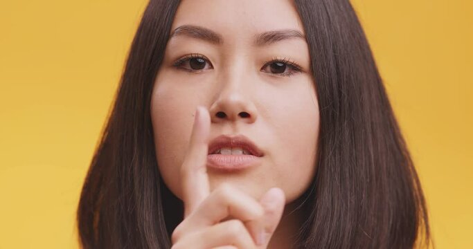 Close up portrait of young asian woman gesturing hush, be quiet, putting finger on lips, orange studio background