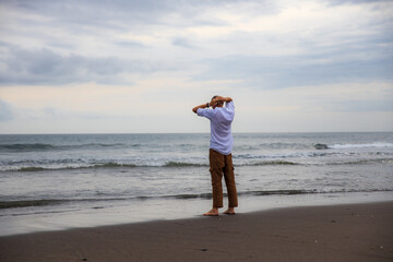 Lonely man standing barefoot on the beach. Man wearing brown trousers and white shirt turned back to the camera. View from back. Horizon seaview. Copy space. Bali, Indonesia