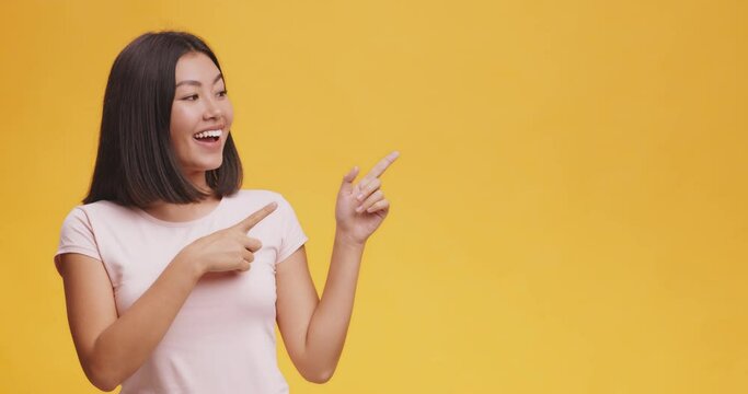 Cheerful asian woman positively pointing aside at copy space, orange studio background, mock up