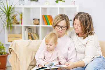 Fototapeta premium Female couple sitting on the sofa of their front room with their child, readding her a book