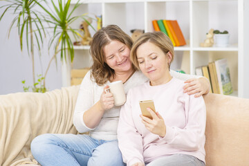 Beautiful young women LGBT lesbian happy couple sitting on sofa using phone and drinking coffee in living room at home. LGBT lesbian couple together indoors concept.  Lesbian couple concept