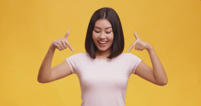 Smiling confident young asian woman pointing fingers on herself, looking at empty space on white t-shirt