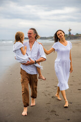 Happy family concept. Summer holidays. Family vacation in Asia. Mother, father and daughter walking barefoot along the beach. Father carrying daughter in his arms. Copy space.
