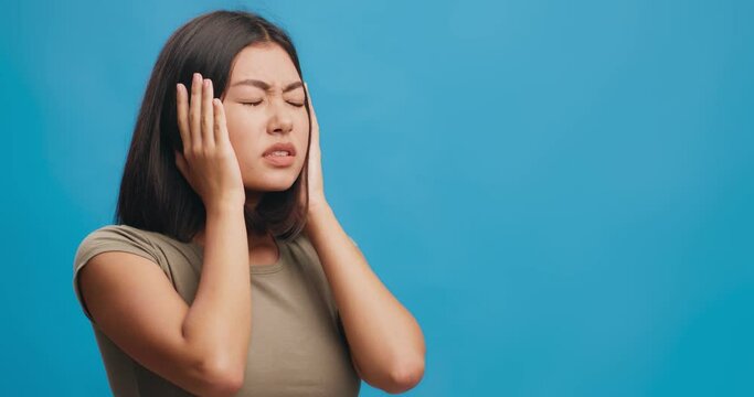 Young asian woman with headache suffering from noise, closing ears with palms, blue background