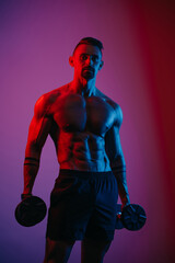 Fototapeta na wymiar A muscular man with a beard who is posing with dumbbells under blue and red lights. An athletic guy is demonstrating his sporty torso. An athlete in shorts after biceps workout.