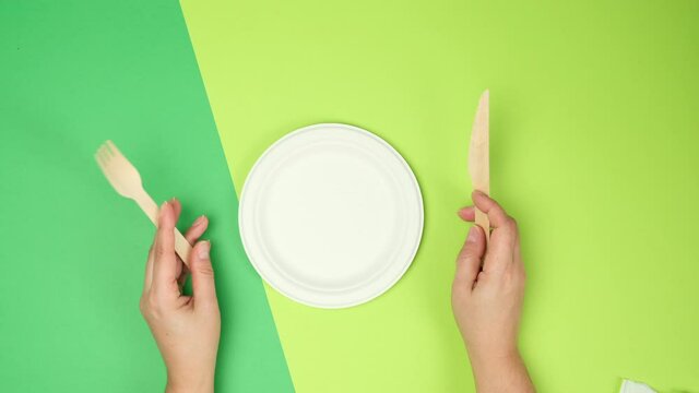 empty white paper plate and female hands are holding disposable fork and knife on a green background, top view