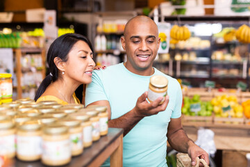 Interested Latin American couple reading product label on jar while choosing groceries in...