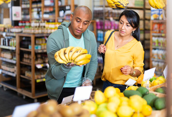 Obraz premium Smiling latin american family couple looking for ripe sweet bananas in food store..