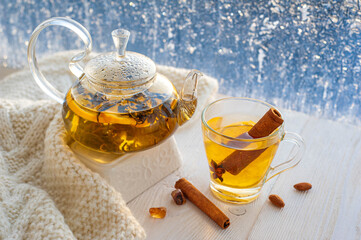 Winter warming tea with lemon and orange against the background of frosty patterns. Hot drink in a glass kettle and a cup on a wooden table in the background of the window.