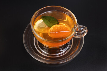 Liquid tea lemon orange slice green leaf cinnamon stick in transparent glass teacup saucer on black background