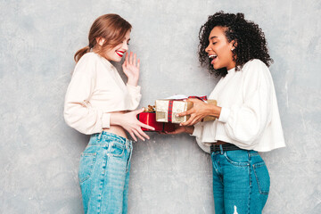 Two young beautiful smiling international hipster female in trendy clothes.Sexy carefree women posing near gray wall.Positive models hugging and giving each other gift boxes.Christmas, x-mas, concept