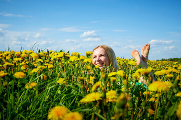 Fototapeta premium Pretty woman on a field with green grass and yellow dandelion flowers in a sunny day. Girl on nature with yellow flowers and blue sky.