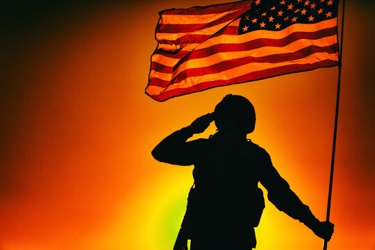 Silhouette of army soldier in combat helmet, armed service rifle, holding USA national flag, saluting on background of sunset or dawn sky. Military respect and honor, patriotism and heroes remembrance