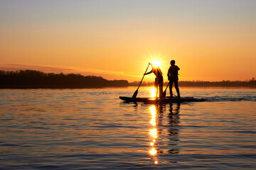 Silhouettes of two boys rowing on supboards (SUP) in winter river at sunset