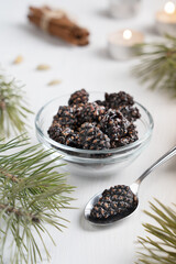 Image with selective focus of siberian healthy dessert of dried pine cones preserved in sweet syrup served in bowl on white wooden background with spoon, pine branches and candle lights. Vertical