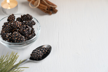 Siberian healthy dessert of dried pine cones preserved in sweet syrup served in bowl on white wooden background with pine branches, spoon, candle light and cinnamon in winter. Image with copy space