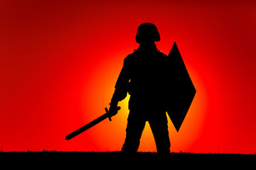 Silhouette of modern soldier in combat helmet, armed sword and shield, standing on background of sunset sky and sun setting behind horizon. Army heroism, state defender and patriotism concept
