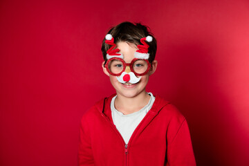 Pretty boy celebrating Christmas wearing funny Christmas glasses in front of red background