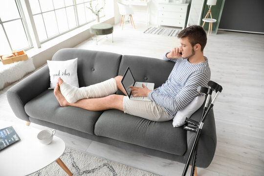 Young man with broken leg using laptop while talking by phone at home