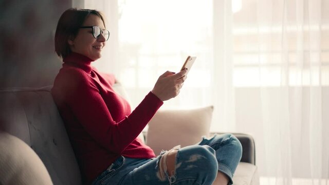 A relaxed young woman in glasses using her smartphone surfing social media, checking the news, playing mobile games, or texting messages while sitting on the sofa.