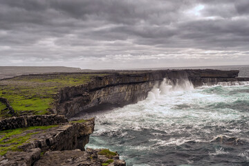 Powerful waves of Atlantic ocean crashes against strong stone cliffs. Landscape of Inishmore Aran islands, county Galway, Ireland. Dark grey sky. Nobody. Nature environment.