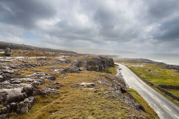Fototapeta na wymiar Landscape in Burren area by Atlantic ocean, West of Ireland. Small narrow asphalt road by the ocean, part of Wild Atlantic Way route. Beautiful cloudy sky. Travel and nature concept.