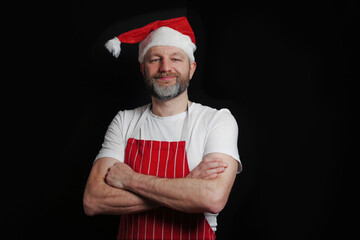 Fototapeta premium Professional butcher or fishmonger in white t shirt and classic apron and Christmas hat. The model is male in his 40s, slim body type, grey beard. Black background, Hands crossed.