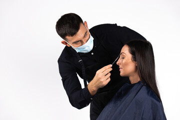 A young man in a medical mask makes a makeup for a women in the studio on a white background. Pandemic beauty work