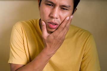 Fototapeta premium A man in yellow T-shirt holding his cheek with hand, suffering from painful toothache. Tooth decay or sensitivity. Dental health and care concept