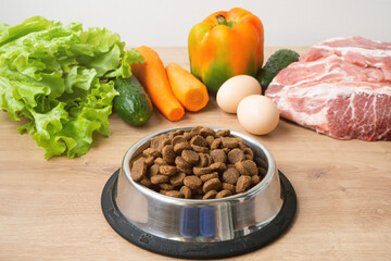 Dry pet dog food with natural ingredients. Raw meat, vegetables, eggs and salad near bowl with dry pet feed on wooden background. concept of pet correct balanced and healthy nutrition