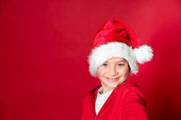 Young pretty boy celebrating Christmas wearing Christmas hat in front of red background