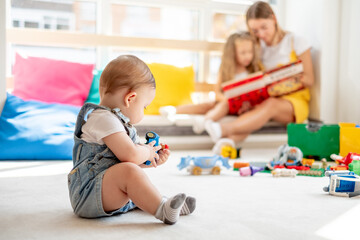 Obraz na płótnie Canvas Baby playing with toys while mother and daughter reading book on background, family time, education