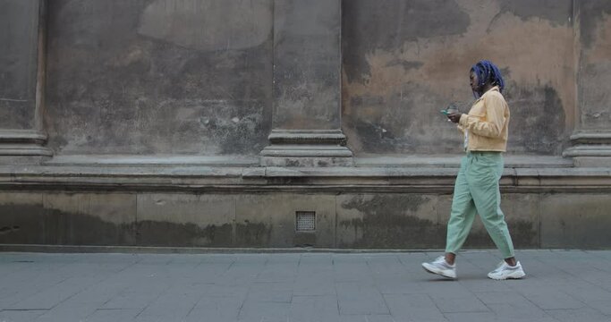 Joyful female person using smartphone and carrying coffee papper cup at city street. Afro american woman with dreadlocks walking and moving in rhytm. Concept of emotions