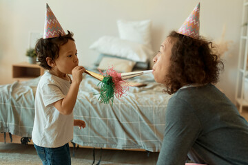 Happy young Hispanic mother with cone hat celebrating third birthday anniversary of her cute little...
