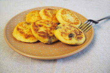 six yellow sugar-free cheesecakes with banana and raisins in cornmeal lie in a brown round polka-dot plate with a metal fork on a gray tablecloth side view . cooking gluten-free cheesecakes at home