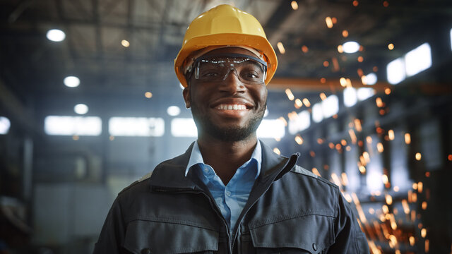 Happy Professional Heavy Industry Engineer/Worker Wearing Uniform, Glasses and Hard Hat in a Steel Factory. Smiling African American Industrial Specialist Standing in a Metal Construction Manufacture.