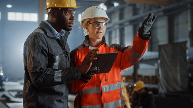 Two Heavy Industry Engineers Stand in Steel Metal Manufacturing Factory, Use Digital Tablet Computer and Have a Discussion. Black African American Industrial Specialist Talk to Female Technician.