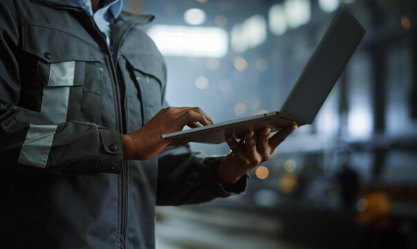 Close Up on Hands of a Professional Heavy Industry Engineer Wearing Safety Uniform and Using Laptop Computer. African American Industrial Specialist Standing in a Metal Construction Manufacture.