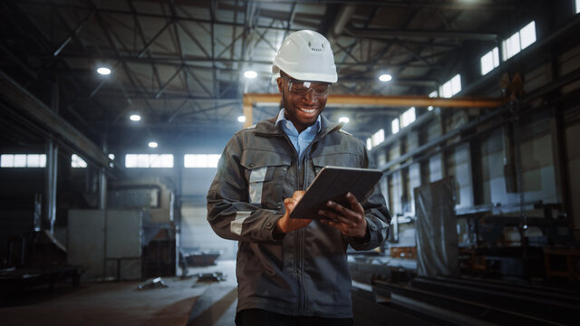 Professional Heavy Industry Engineer/Worker Wearing Safety Uniform And Hard Hat Uses Tablet Computer. Smiling African American Industrial Specialist Walking In A Metal Construction Manufacture.