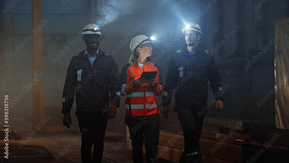 Aufkleber Three Diverse Multicultural Heavy Industry Engineers and Workers in Uniform Walk in Dark Steel Factory Using Flashlights on Their Hard Hats. Female Industrial Contractor is Using a Tablet Computer.