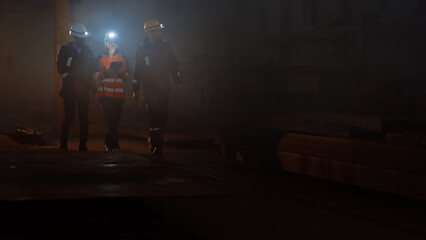 Three Diverse Multicultural Heavy Industry Engineers and Workers in Uniform Walk in Dark Steel Factory Using Flashlights on Their Hard Hats. Female Industrial Contractor is Using a Tablet Computer.