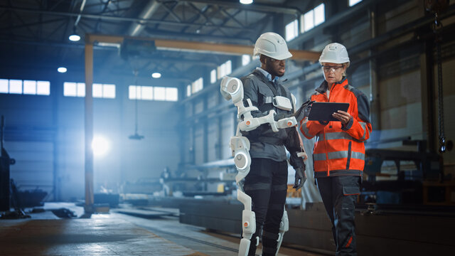 Futuristic Concept of a Manual Labor Worker in a Bionic Exoskeleton Prototype Working in a Factory. Heavy Industry Engineer Monitors the Powered Suit on an African American Assistant.