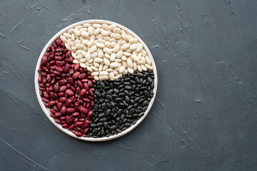 beans in white ceramic plate. bowl of beans. black bean, red kidney bean and white bean. navy, cannellini, white kidney bean on dark gray background top view photo