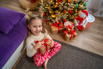 Happy smiling baby girl with deer antlers holds a gift box near Christmas tree