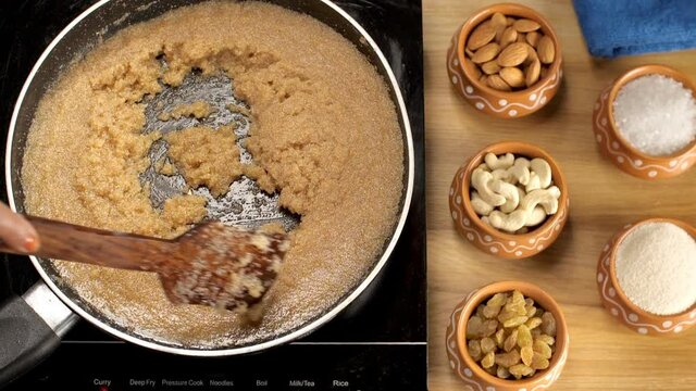 Preparation of Indian sweet/dessert - Sooji/Suji/semolina Halwa in Desi Ghee. Lady's hand stirring a pan full of Sooji Ka Halwa with almonds  cashew nuts  raisins  sugar kept at the side - Indian c...