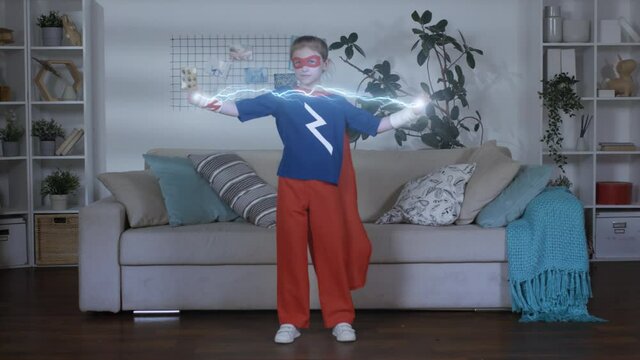 Full shot of young girl in superhero costume with cloak and mask standing in room controlling lightning bolt in hands suddenly clapping so lightning disappearing