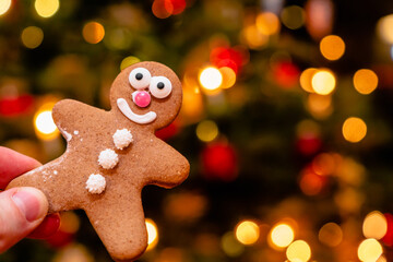 Hand holding a smiling gingerbread man in front of a blurred background with warm bokeh lights. Christmas lights background. Celebrating winter holidays, advent, Christmas season. Festive decoration.